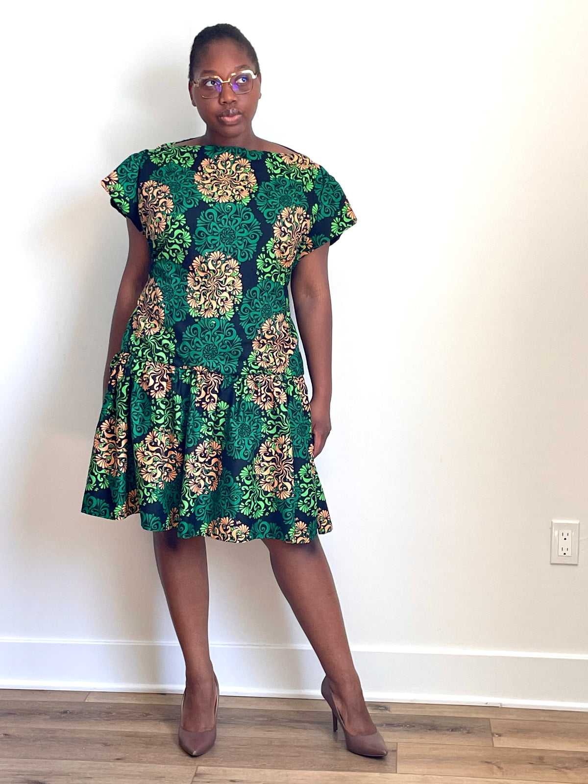 A woman modeling a short-sleeve knee-length dress with a green, gold, and beige floral pattern, standing against a plain white wall on a light wood floor.