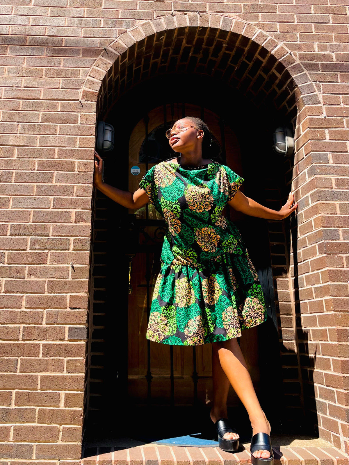 A woman wearing a green and gold patterned dress stands in the doorway of a brown brick building, posing with her hands on the brick arch.