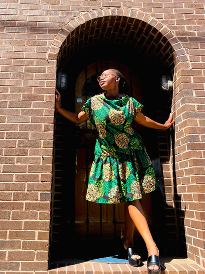 A woman wearing a green and gold patterned dress stands in the doorway of a brown brick building, posing with her hands on the brick arch.