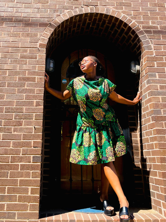 A woman wearing a green and gold patterned dress stands in the doorway of a brown brick building, posing with her hands on the brick arch.