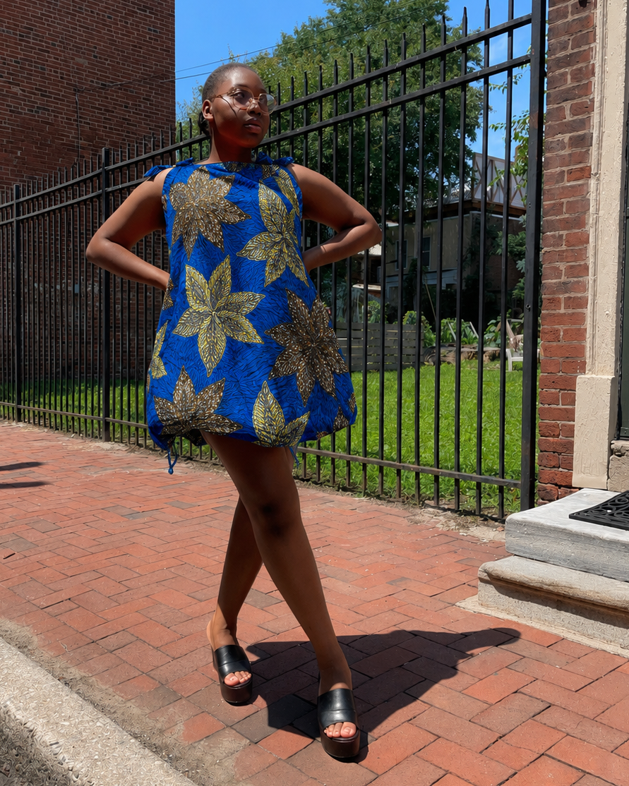 A woman wearing a blue sleeveless dress with a gold floral pattern stands on a red brick sidewalk outdoors, posing with her hands on her hips. The background features a black metal fence, a brick building, and green grass under a blue sky.